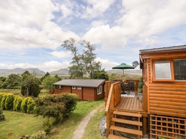 A wooden cabin with outdoor seating and umbrella surrounded by grass and trees at Ty Coed - 63 in Bronaber near Trawsfynydd
