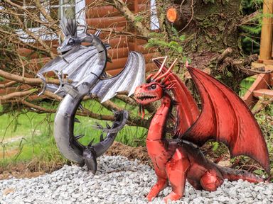 Metal sculptures of a silver dragon and a red dragon on gravel near a tree outdoors