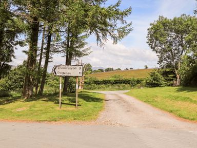 A rural road with trees and a roadside sign showing distances to Trawsfynydd and Dolgellau at Ty Coed - 63 in Bronaber near Trawsfynydd