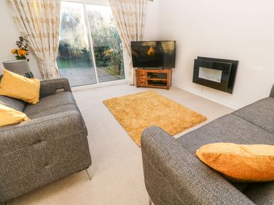 A living room with two grey sofas and yellow cushions a TV on a wooden stand a wall-mounted fireplace and sliding glass doors with curtains at Dunes View in Cubert