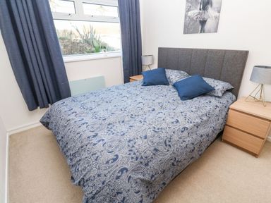 A bedroom with a double bed with patterned bedspread and two blue pillows two wooden bedside tables with lamps and a window with blue curtains at Dunes View in Cubert