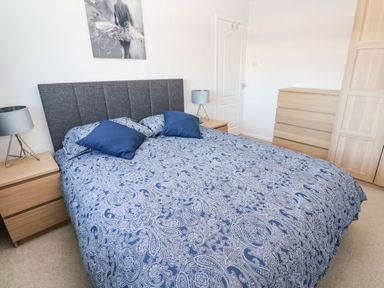 A bedroom with a bed with patterned bedding and two blue pillows two bedside tables with lamps a wooden chest of drawers and a wardrobe at Dunes View in Cubert