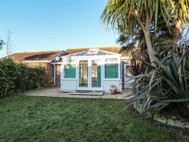 A backyard with grass and a tiled patio leading to a conservatory attached to a brick house at Dunes View in Cubert