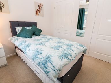 A bedroom with a single bed with palm tree patterned bedding two green pillows a bedside table a wall mirror and white double wardrobes at Dunes View in Cubert