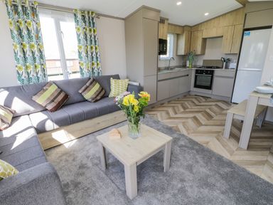 A living room with a grey sofa and a small wooden coffee table with flowers in a vase next to a kitchen with wooden cabinets at Jeans Place in East Heslerton near West Heslerton