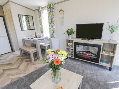 A living room with flowers on a coffee table a dining table set with plates and glasses a television on a stand with a fireplace below at Jeans Place in East Heslerton near West Heslerton