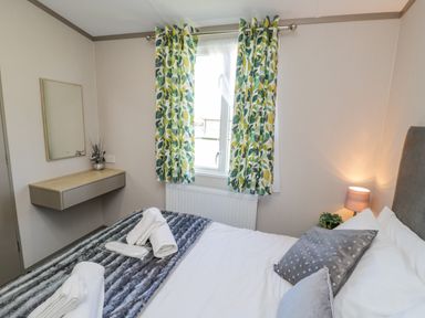 A bedroom with a bed, pillows, folded towels, a bedside lamp, a wall mirror, a floating desk, and a window with patterned curtains at Jeans Place East Heslerton near West Heslerton