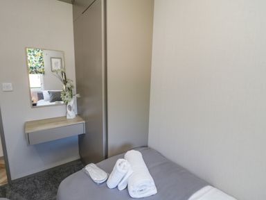 A small bedroom with a bed covered in gray fabric with folded white towels and slippers on top and a wall-mounted wooden shelf with a vase and a mirror reflecting another room at Jean's Place in East Heslerton near West Heslerton