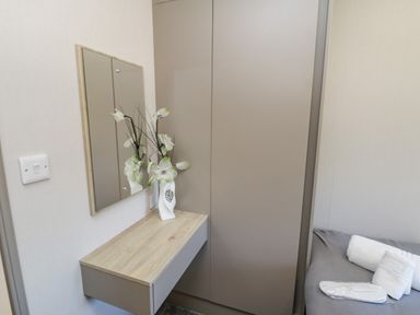 A corner of a bedroom with a wall-mounted wooden vanity table, a vase with white flowers, a mirror, a closet, and a bed with towels and slippers at Jeans Place in East Heslerton near West Heslerton