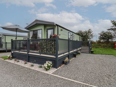 A green mobile home with a black wooden deck and railing surrounded by gravel and flowers at Jean's Place in East Heslerton near West Heslerton