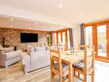 A dining table with wooden chairs and cushioned seats in an open living area with sofas and a wall-mounted tv at Little Lodge in Pentney near King's Lynn
