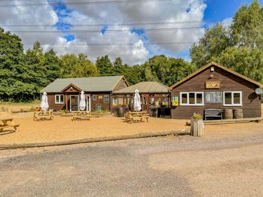 An outdoor dining area with picnic tables and umbrellas in front of a wooden cafe building surrounded by trees at Little Lodge in Pentney near King's Lynn