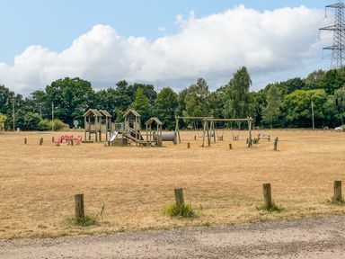 A playground with swings slides and climbing structures in a large open field near trees and a power line at Little Lodge in Pentney near King's Lynn