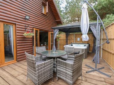 An outdoor wooden deck with a glass table and four wicker chairs a hot tub under a gazebo and a closed umbrella at Little Lodge in Pentney near King's Lynn
