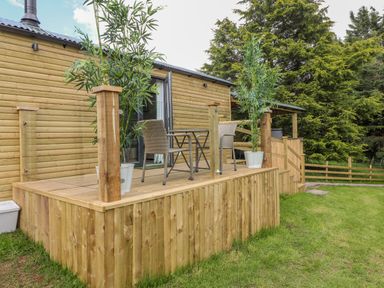 An outdoor wooden deck with two wicker chairs and a table in front of a wooden cabin at High Rigg Shepherd's Sunrise in Brampton Cumbria