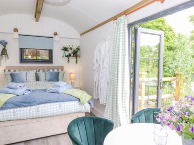 A bedroom with a bed and blue and green bedding next to a table with flowers and chairs by an open door showing greenery at High Rigg Shepherd's Sunrise in Brampton Cumbria