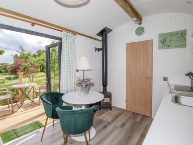 A small dining area with a round white table and green chairs near a wood stove and open door leading to a wooden deck at High Rigg Shepherd's Sunrise in Brampton Cumbria