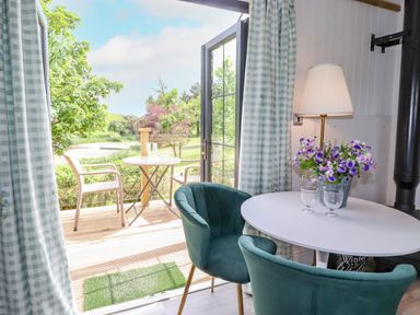 A small dining area with two green chairs and a white table with flowers next to an open door leading to a patio with a table and two chairs at High Rigg Shepherd's Sunrise in Brampton Cumbria