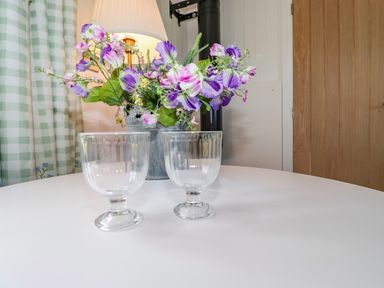 A white round table with two clear glass goblets a metal vase with purple and pink flowers a lamp green checkered curtains and a wooden door in a room at High Rigg Shepherd's Sunrise in Brampton Cumbria