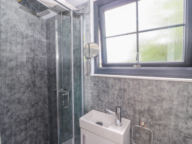 A bathroom with a shower enclosure a small white sink a round magnifying mirror and a window at High Rigg Shepherd's Sunrise in Brampton Cumbria
