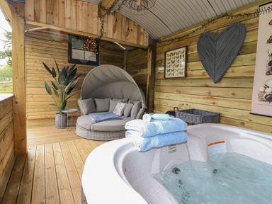 A wooden outdoor room with a hot tub towels and a round cushioned chair with pillows at High Rigg Shepherd's Sunrise in Brampton Cumbria