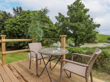 An outdoor wooden deck with a round glass top table and two woven chairs overlooking a pond and trees at High Rigg Shepherd's Sunrise in Brampton Cumbria