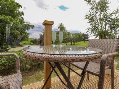 An outdoor seating area with a round glass table and two wicker chairs overlooking a grassy field at High Rigg Shepherd's Sunrise in Brampton Cumbria