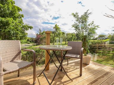 A wooden deck with a round glass table and two wicker chairs overlooking a green garden at High Rigg Shepherd's Sunrise in Brampton Cumbria