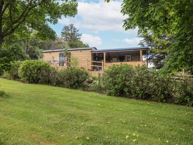 A wooden cabin with a deck and outdoor furniture surrounded by trees and bushes at High Rigg Shepherd's Sunrise in Brampton Cumbria