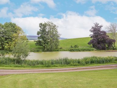 A pond surrounded by grass and trees with hills and blue sky in the background at High Rigg Shepherd's Sunrise in Brampton Cumbria