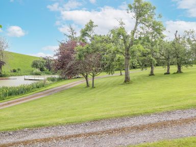 A grassy area with trees a gravel driveway and a small pond with a wooden dock at High Rigg Shepherd's Sunrise in Brampton Cumbria