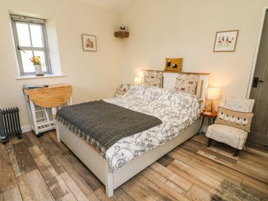 A bedroom with a bed and patterned bedding a chair with a cushion and a small wooden table with a lamp at Herdie Hideaway in Appleby-In-Westmorland