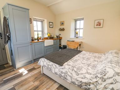 A bedroom with a bed a small kitchenette with blue cabinets and a wooden drop leaf table near two windows at Herdie Hideaway in Appleby-In-Westmorland