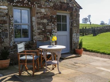 An outdoor stone patio with a small round table and two wooden chairs beside a stone building with a window and door at Herdie Hideaway in Appleby-In-Westmorland