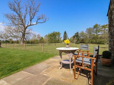 An outdoor patio with two wooden chairs a small round table with flowers and a grassy field with trees at Herdie Hideaway in Appleby-In-Westmorland