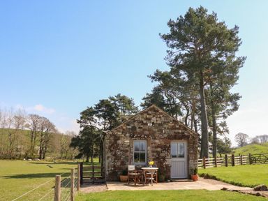 A stone cottage with a small patio table and chairs outside surrounded by a grassy field and trees at Herdie Hideaway in Appleby-In-Westmorland