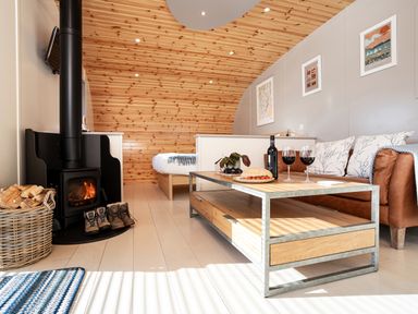 A small living area with a wood stove fireplace boots and firewood next to a coffee table with wine and food and a sofa and bed in the background at Middle Longlands in Cartmel