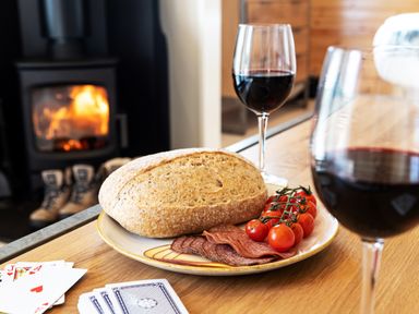 A plate with bread slices salami and cherry tomatoes on a wooden table with two glasses of red wine and playing cards near a fireplace at Middle Longlands in Cartmel