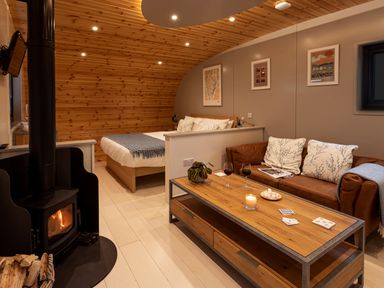 A room with a bed separated by a half wall from a seating area with a sofa and a wooden coffee table with a wood stove at Middle Longlands in Cartmel