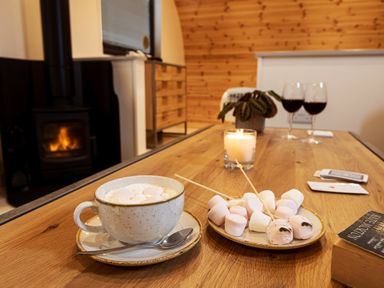 A wooden table with a cup of hot chocolate with marshmallows a plate of marshmallows a lit candle two glasses of red wine and a book by a fireplace at Middle Longlands in Cartmel