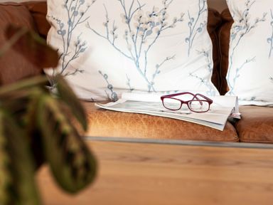 A leather sofa with white cushions with blue floral patterns and a magazine with red reading glasses on it
