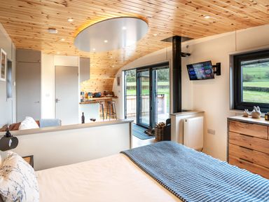 A bedroom with a bed and a blue blanket next to a wooden dresser and a mounted TV with a kitchen and sliding glass doors in the background at Middle Longlands in Cartmel