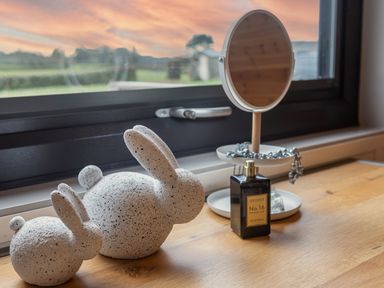 A wooden table by a window with two speckled stone bunny sculptures a round mirror with a jewelry tray and a perfume bottle at Middle Longlands in Cartmel