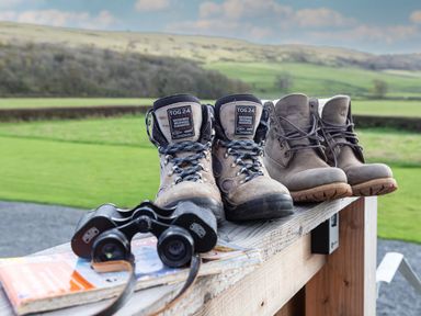 Two pairs of walking boots next to binoculars and a map on a wooden surface with fields and hills in the background at Middle Longlands in Cartmel