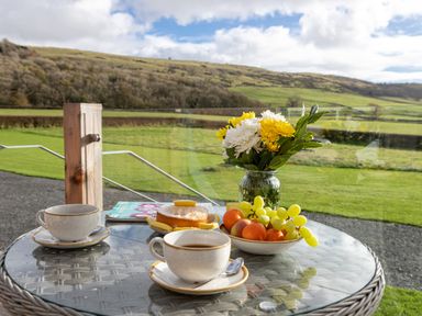 A glass table with cups of coffee a cake a bowl of fruit and a vase of flowers on a patio overlooking green fields at Middle Longlands in Cartmel