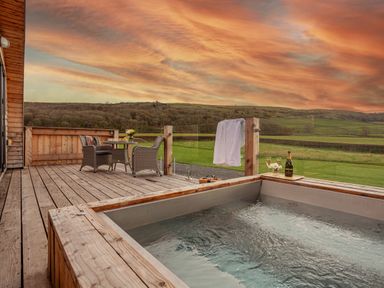 An outdoor wooden deck with a hot tub a table with chairs and champagne with glasses at Middle Longlands in Cartmel