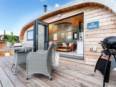 An outdoor deck with wicker chairs table and a barbecue outside a wooden pod at High Longlands in Cartmel