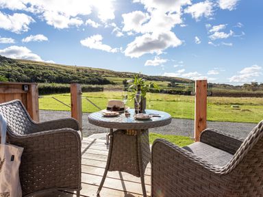 An outdoor seating area with two woven chairs and a glass top table with drinks and snacks overlooking grassy fields at High Longlands in Cartmel