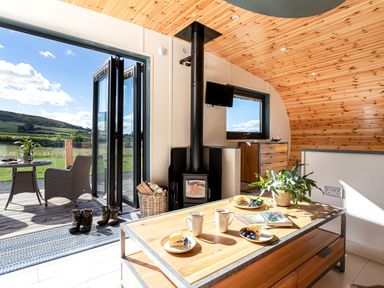 An interior living space with a wood stove table with cups plates and a plant open doors to a deck with chairs and a table at High Longlands in Cartmel