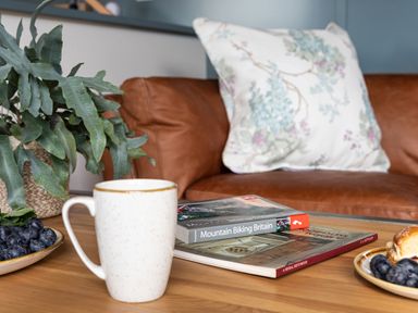 A coffee table with a white speckled mug a plant two books and plates of blueberries in front of a brown leather sofa with a floral pillow at High Longlands in Cartmel